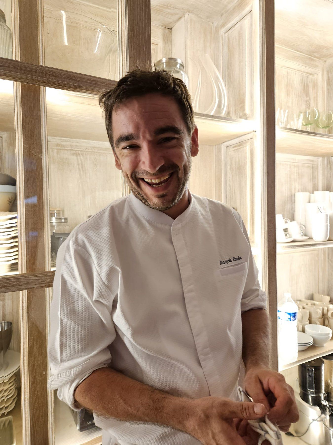 Chef François Savin in his whites, laughing in front of a dresser filled with porcelain, a service cloth in his hands.
