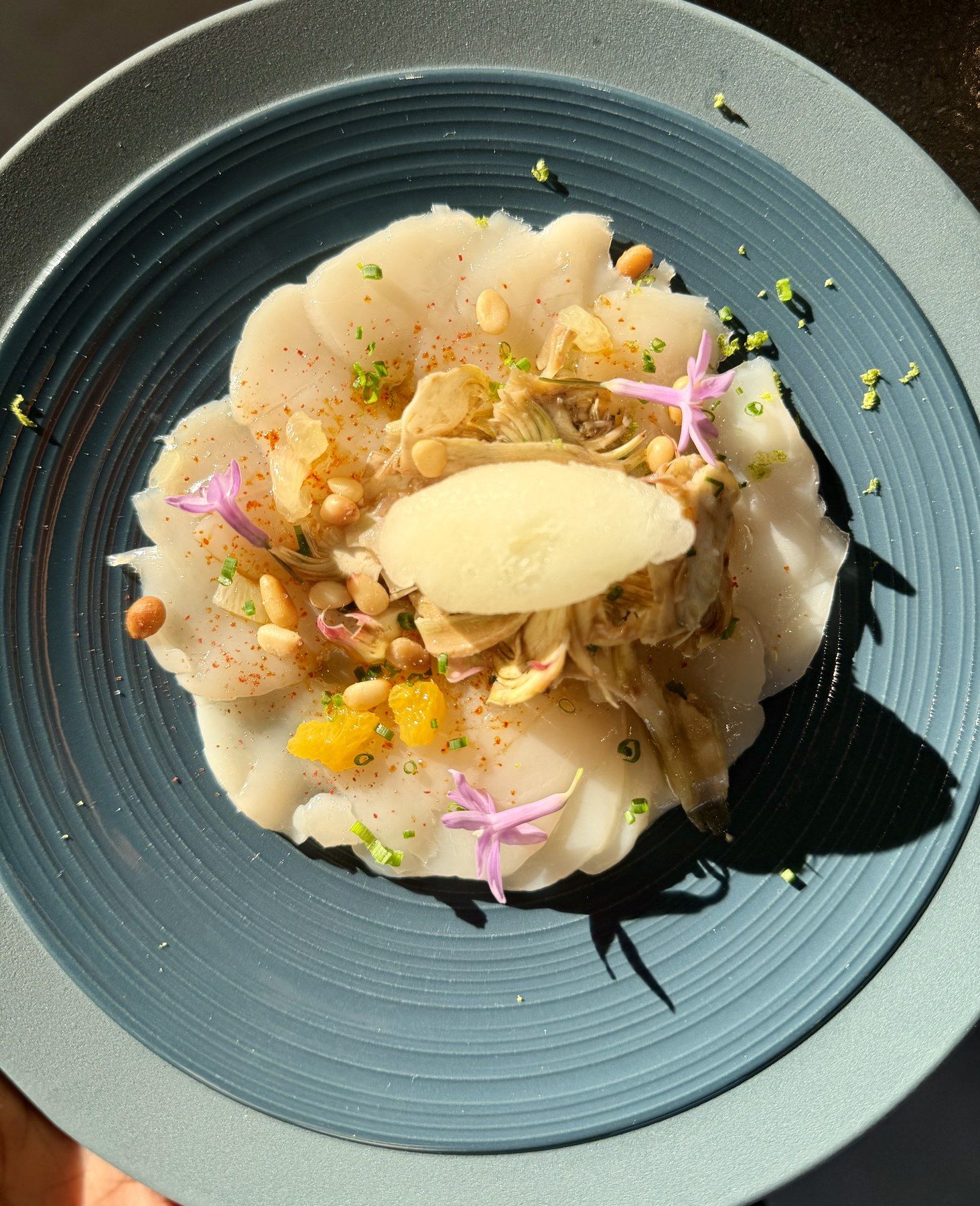 Scallop carpaccio with artichoke, pine nuts and edible flowers on a blue plate.