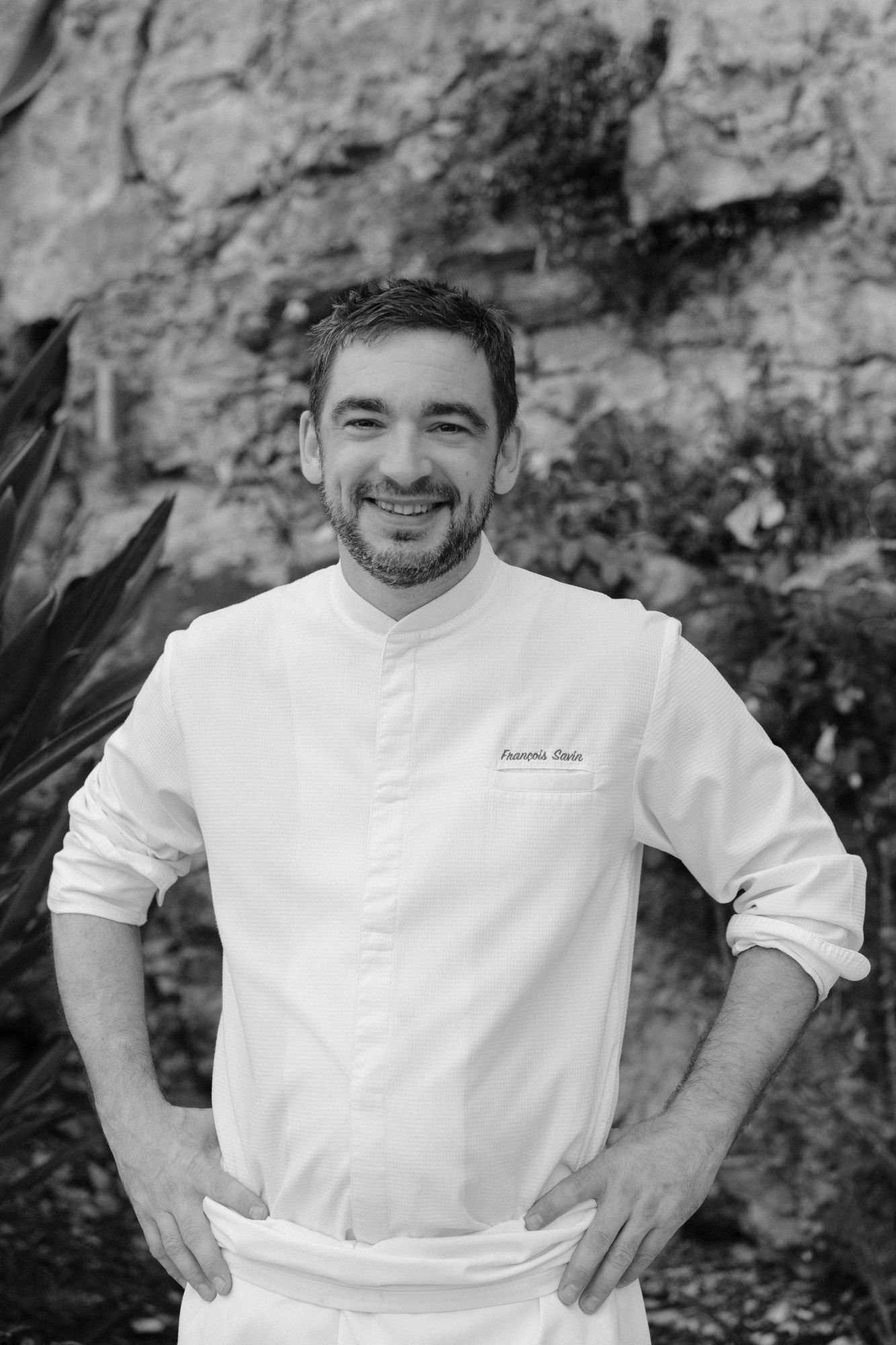 Chef François Savin in his whites, smiling, an outdoor portrait against a Provençal stone wall.