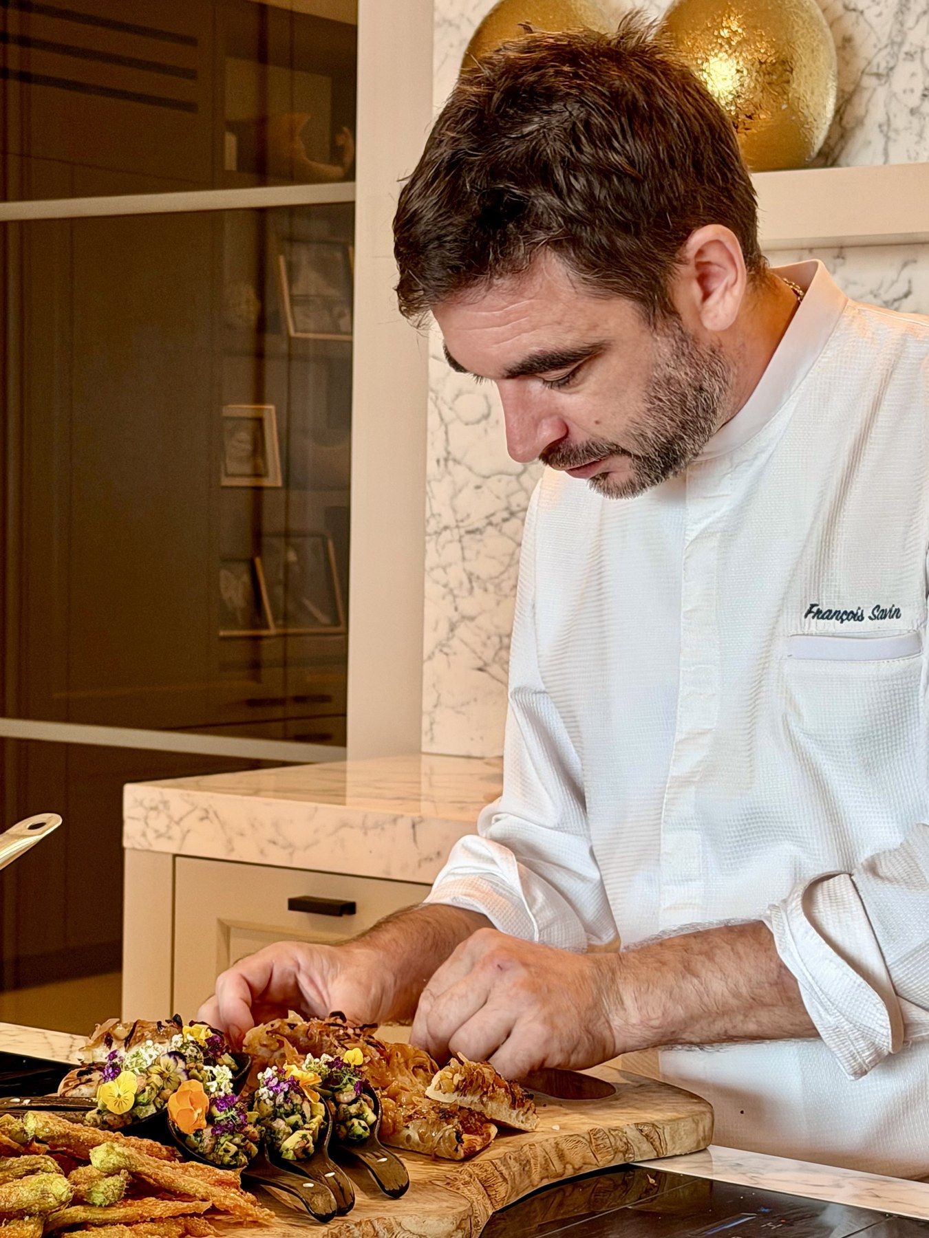 The chef plating amuse-bouche spoons on an olive-wood board.
