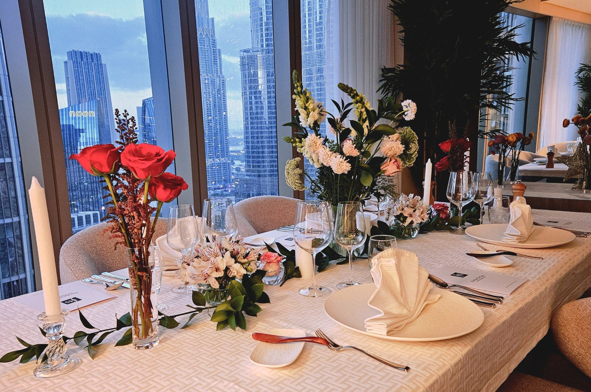 A dinner table dressed in white linen and flowers, set for an evening before the lit Dubai skyline.