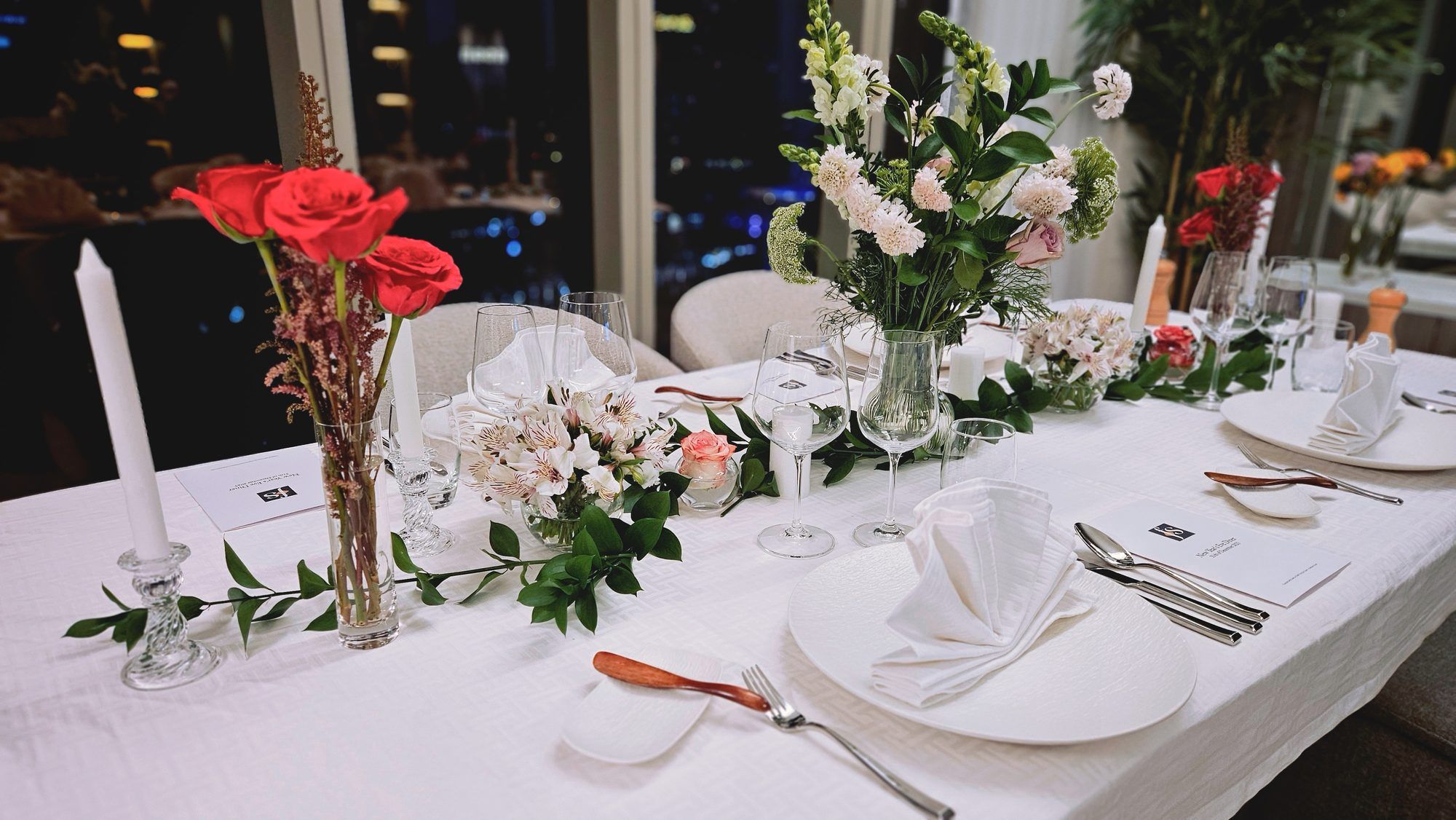 A long table dressed in white linen with red roses and white blooms, set for an evening dinner before floor-to-ceiling windows overlooking the Dubai skyline at night.