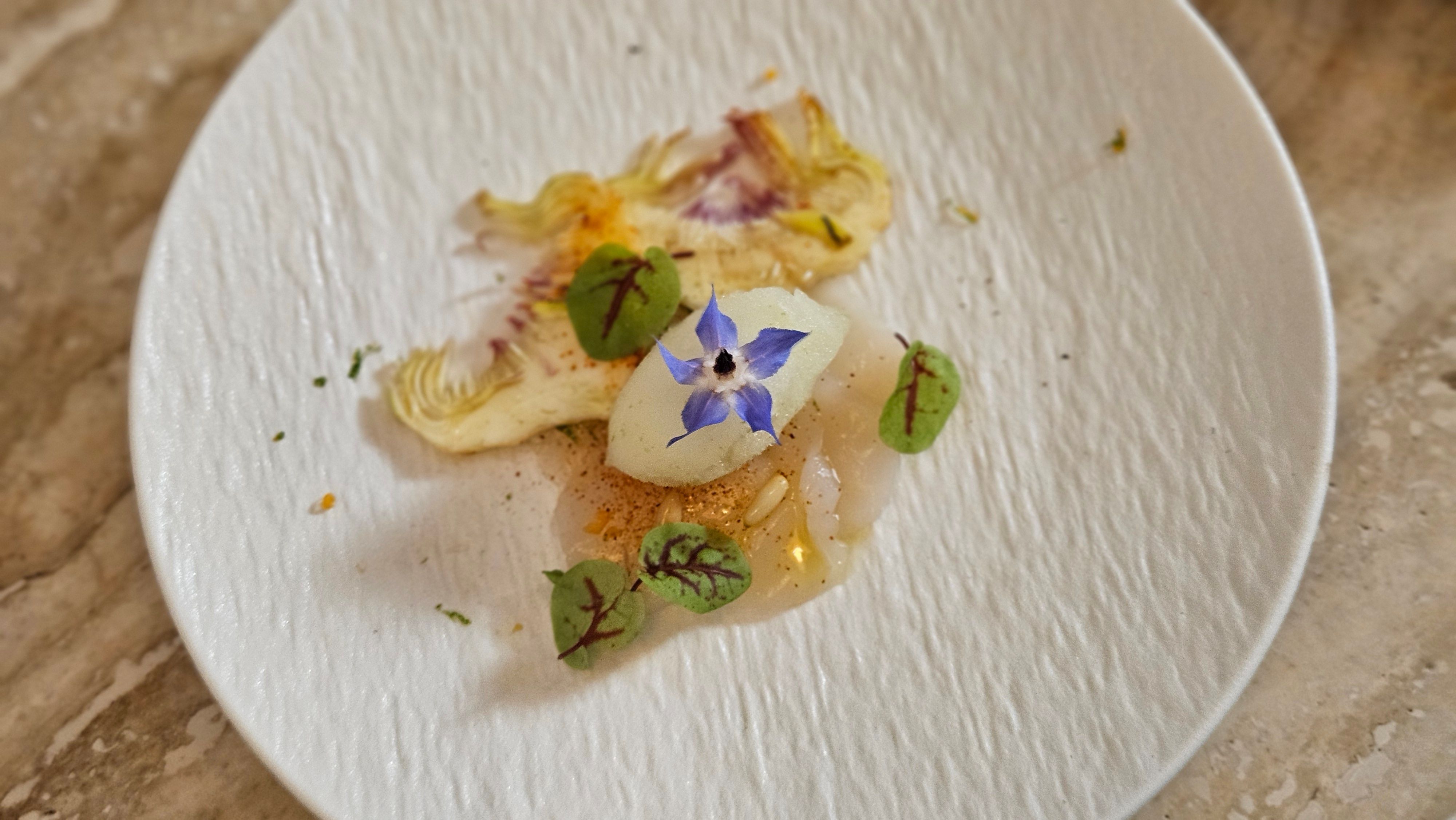 King scallop carpaccio with shaved artichoke, a borage flower, pine nuts and oxalis on a white textured plate.