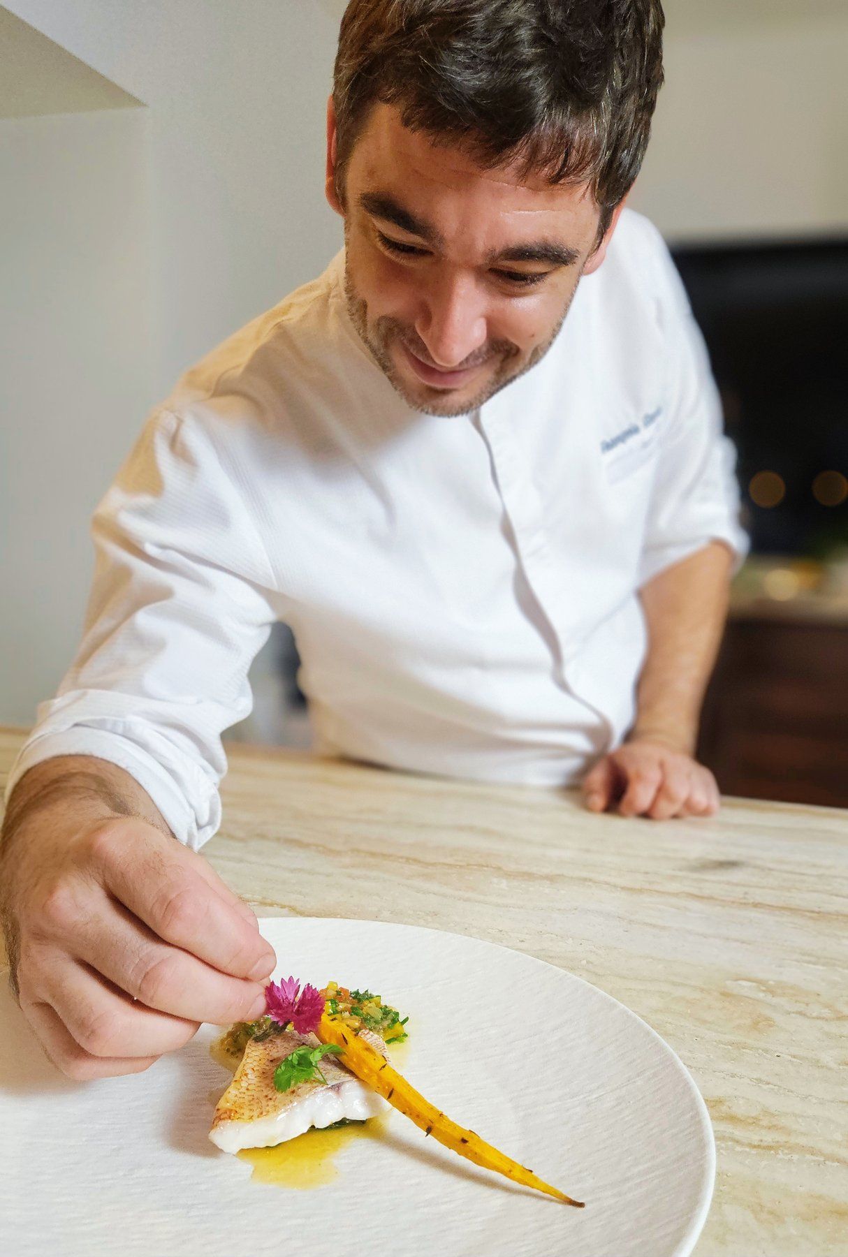 Chef François Savin in his whites, plating a fish fillet with a glazed baby carrot and herb garnish.