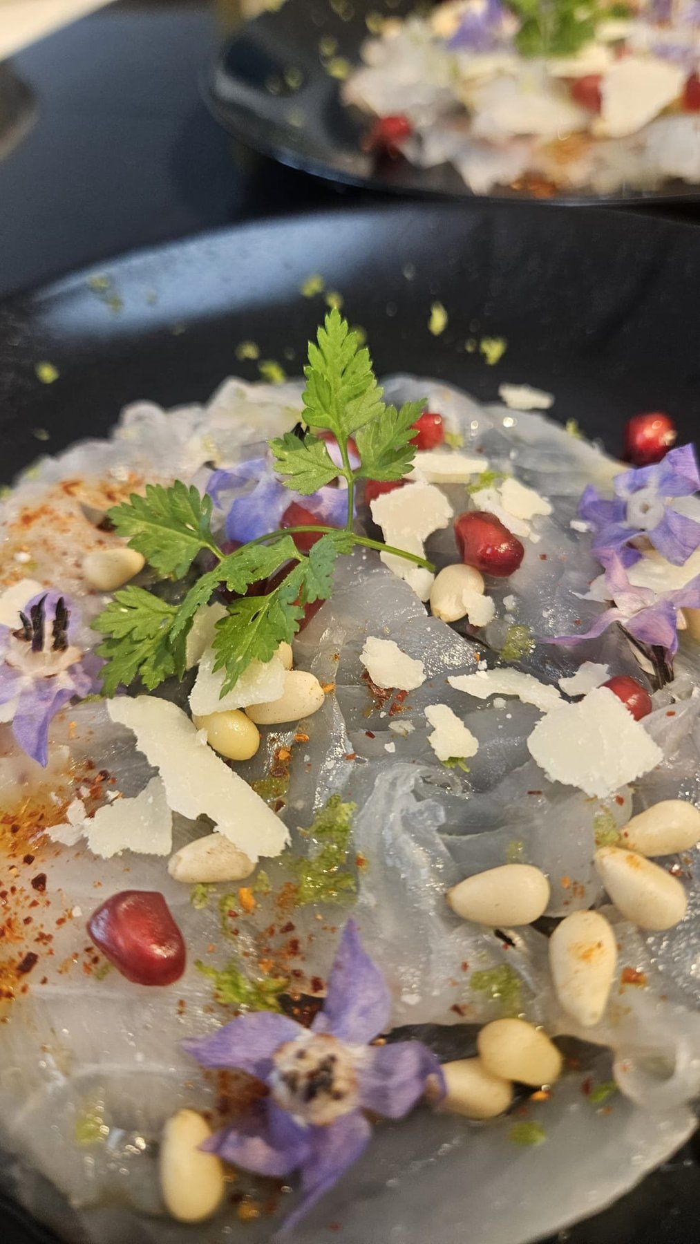 Sea-bream carpaccio on a dark plate with pomegranate, shaved parmesan, pine nuts and edible flowers.
